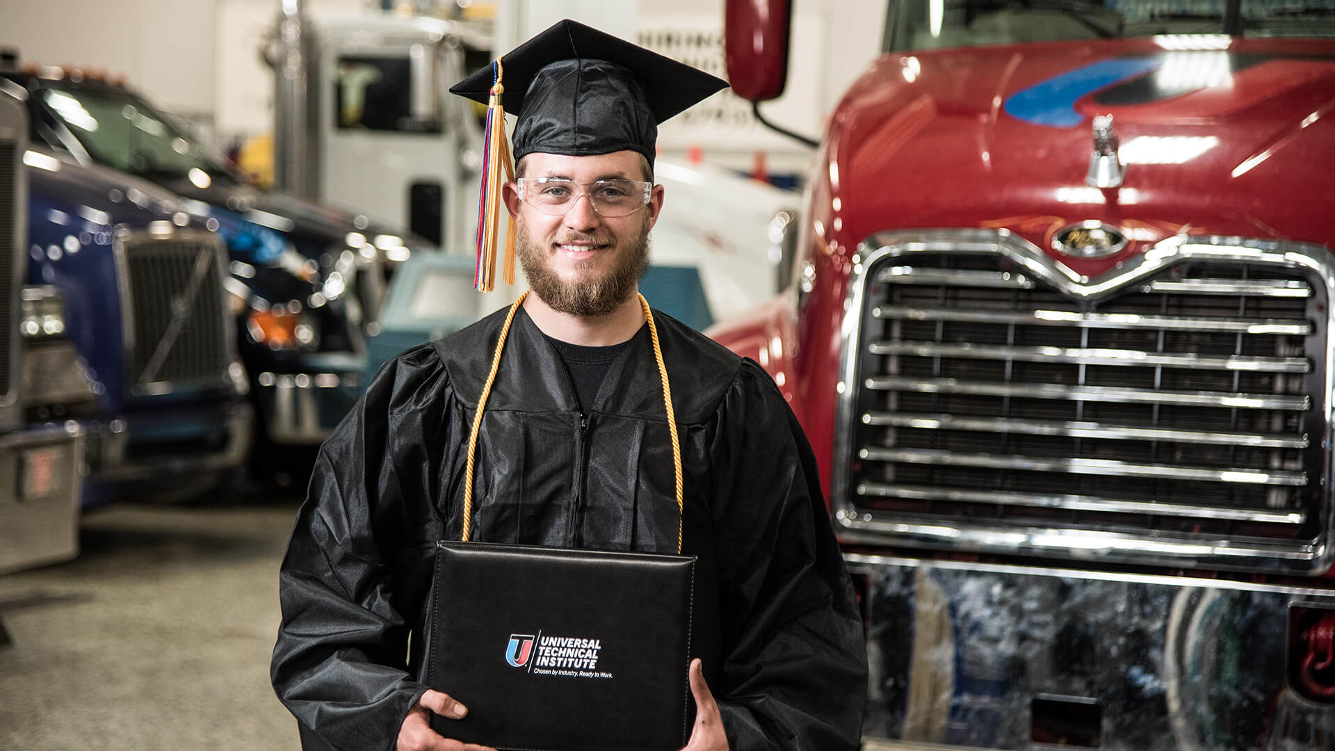 UTI student in a cap and gown holding a diploma smiling with diesel trucking in the background